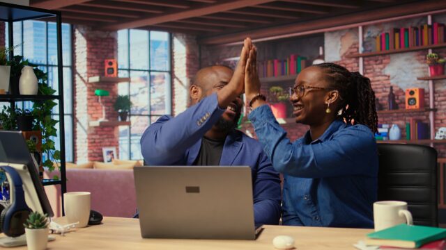 Black freelancing couple doing a high five to celebrate their achievement, completing tasks to ensure successful project. Young man and woman feeling pleased with their cooperation. Camera A.