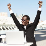 Hurrah! Successful businessman in formal clothes raising his hands having cheerful expression after signing contract. Dark-skinned male working with laptop while sitting outdoors at white table