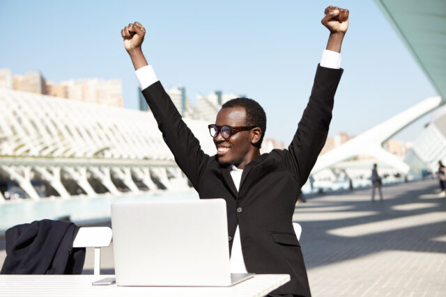 Hurrah! Successful businessman in formal clothes raising his hands having cheerful expression after signing contract. Dark-skinned male working with laptop while sitting outdoors at white table