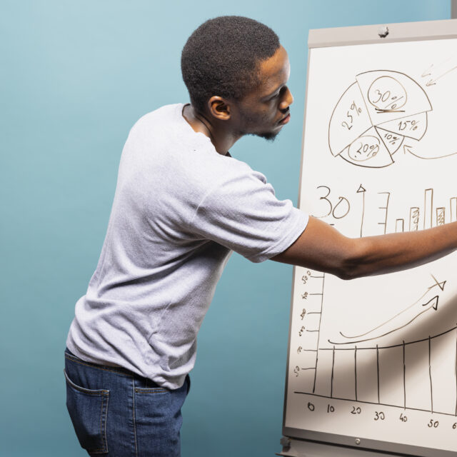 African american male professional in casual clothing drawing graphs and charts on a sketch board. Black man standing next to whiteboard and preparing data for presentation in blue background studio.